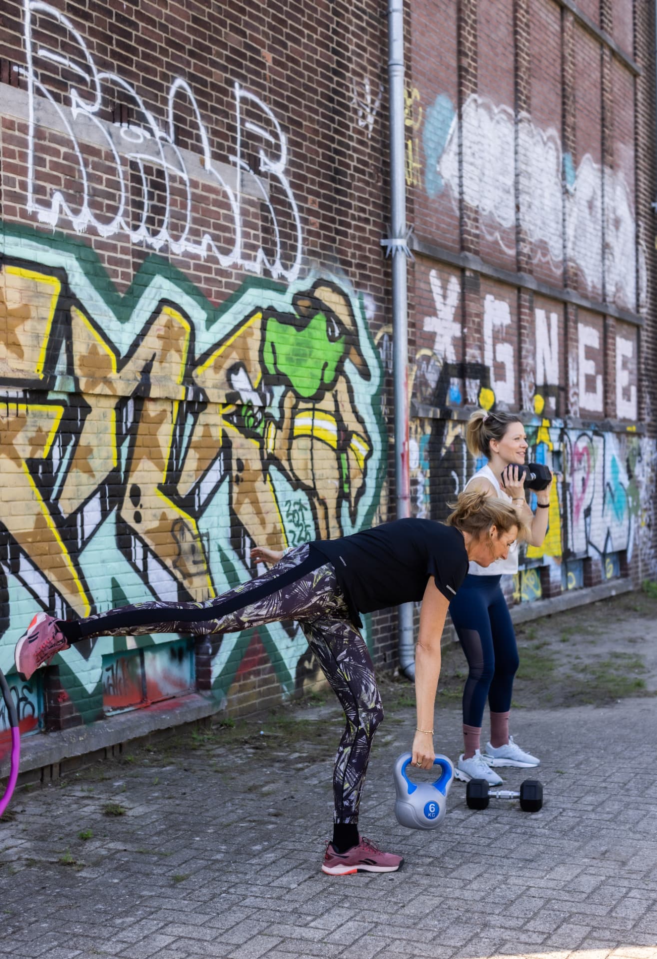 Twee collega's trainen buiten met kettlebell en dumbbell bij een graffitimuur